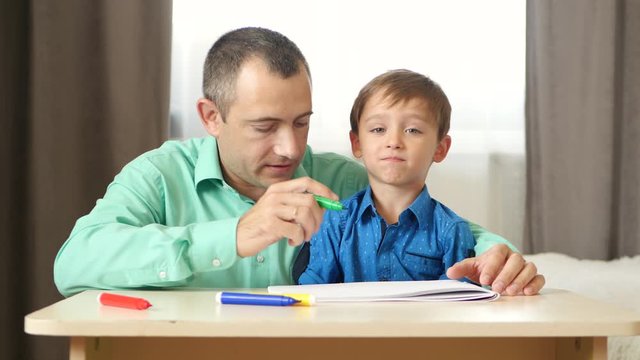 A happy father spends time with his son teaching and drawing with him. Dad and child draw with colored pencils, sitting at the table.