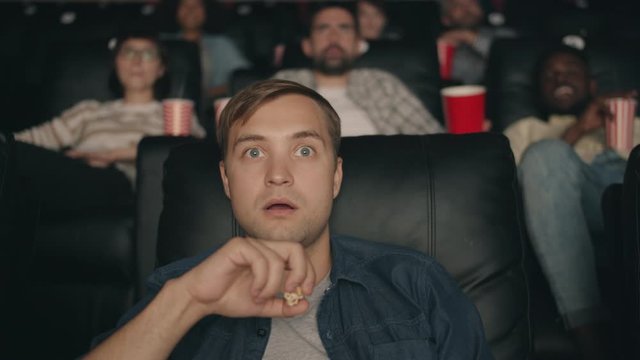 Shocked student blond guy is watching film in cinema with open mouth then eating popcorn smiling enjoying video. Snacks, emotions and entertainment concept.