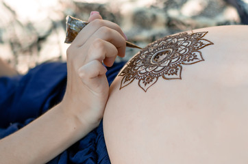 Master is drawing henna tatto on pregnant belly, closeup.  A mehendi artist paints a beautiful...