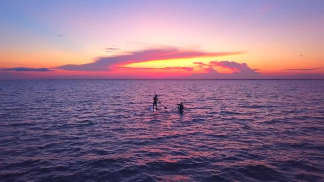 A dramatic drone shot of a kayaker and padlle boarder slowly driffting into a colorful sunset