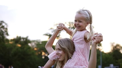 Happy caucasian mother carries beautiful little daughter on her shoulders, dancing and having fun together in public, green park. Day off, motherhood