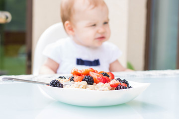 White plate bowl with oatmeal with blackberries and strawberries on a white table. Side view. In the background, a blurry baby is out of focus. Breakfast for children, vegetarian food