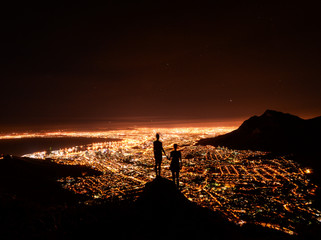Two friends holding hands looking over Cape Town city lights from on top of Lion's Head at night,...