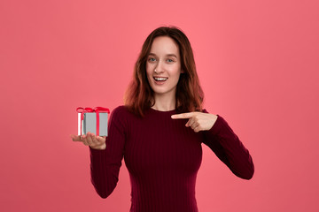 Excited pretty brunette girl holding present box with a ribbon standing isolated on a dark pink background and smiling at the camera. Celebrating special event.