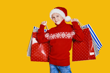 Cute boy in red sweater holding shopping bags with Christmas gifts and looking at camera on yellow background