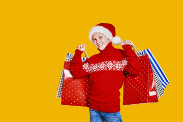 Cute boy in red sweater holding shopping bags with Christmas gifts and looking at camera on yellow background