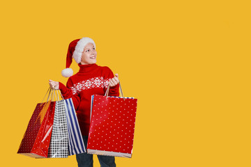 Cute boy in red sweater holding shopping bags with Christmas gifts and looking side away from camera on yellow background