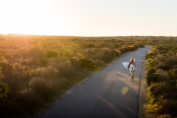 Beautiful blonde surfer girl on her way to the beach on her bicycle with her surfboard.
