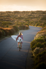 Beautiful blonde surfer girl on her way to the beach on her bicycle with her surfboard.