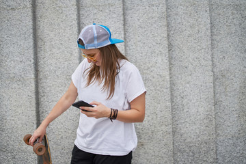 Image of happy female athlete in sunglasses with phone in hand standing with skateboard against gray granite wall
