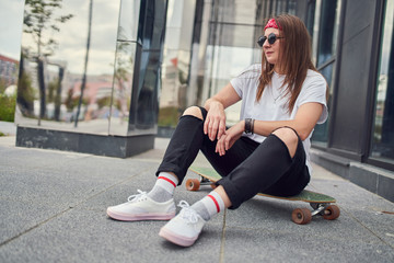 Photo of brunette in sunglasses sitting on skateboard on background of modern buildings in city