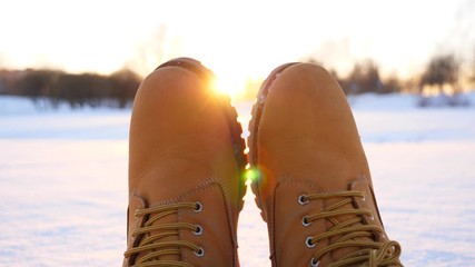 Warm yellow boots against evening sun, man relaxing sitting on snow, enjoy nice winter weather. Bright star of sunlight shine ahead, blurred city park on background. POV slow motion shot