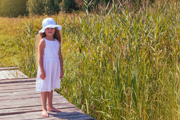 A child in a hat in sunny weather stands on a bridge by a pond