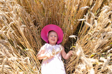 A child, a girl in a pink wide-brimmed hat, lies on a field with ears of rye. Autumn theme, village and people.