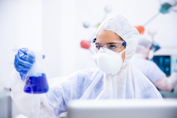 Chemist in goggles and gloves mask holds a container with blue liquid.