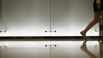 Woman walk with trolley case against glass wall, low shot of legs and rolling silver bag. Lady wear white shirt and short skirt, calmly go at modern hall