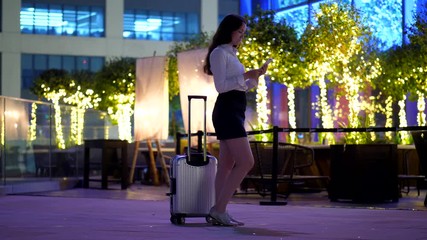 Woman stay still with suitcase at roof terrace, read from smartphone, late evening time. Lady focused on mobile device, bright garland and bushes on background