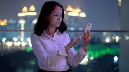 Young adult business woman type text on smartphone, half length portrait shot at rooftop terrace, dim blurred city lights on background. Lady look to phone display and write message, using popular app