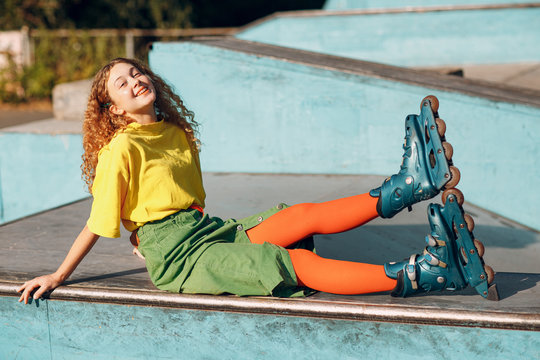 Young woman girl in green and yellow clothes and orange stockings with curly hairstyle roller skating sitting in skate park
