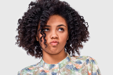 Close-up portrait of confused African American young woman student has dilemma expression, frowns her face and looking up. Doubtful dark skinned female can`t make choice, isolated on white wall