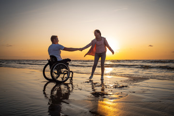 Handicapped man in wheelchair and his girlfriend on a beach at sunset
