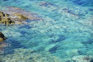 Fototapeta na wymiar Turquoise sea water with grey natural stones. Purity ripple ocean water surface with selective focus and sun light. Transparent blue water surface with rocks. Caribbean water background. Halkidiki