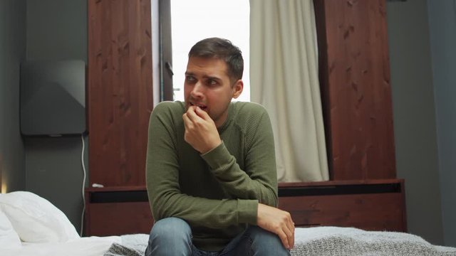 Depressed young man sitting on bed in hotel room and biting nails in despair. Man worrying so much in anxiety or suffering from substance abuse