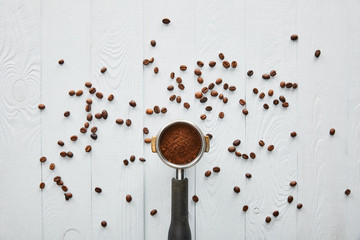 Top view of portafilter with coffee on white wooden surface with scattered coffee beans
