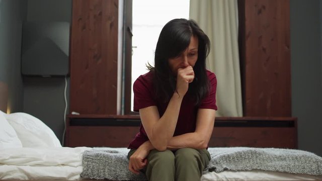 Depressed mature woman sitting on edge of bed at home or in hotel room and suffering from anxiety. Nervous woman worrying so much