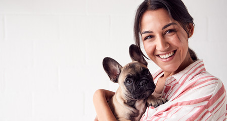 Studio Portrait Of Smiling Young Woman Holding Affectionate Pet French Bulldog Puppy