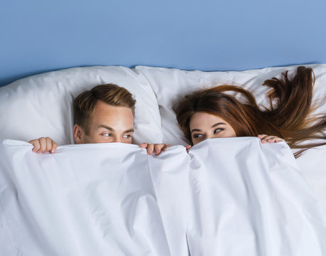 Portrait picture of young playfull couple peeping from bedsheet on the bed at bedroom.
