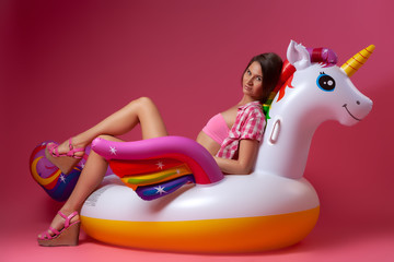 The concept of cheerful bright mood and fun. Bright portrait of a young woman in shorts, a pink top and a shirt  resting on an inflatable unicorn mattress on an isolated pin k background