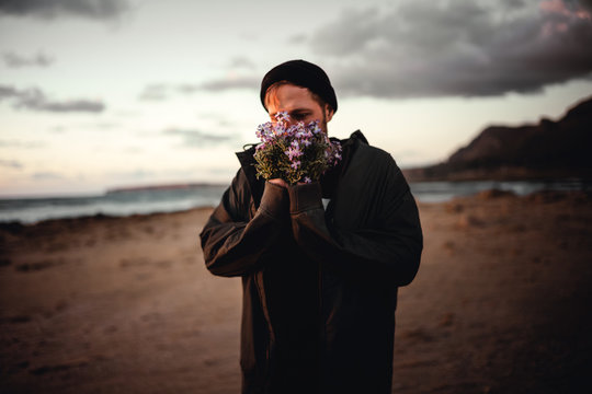 Junger Mann mit Jacke und M&uuml;tze steht mit Wild Blumen Strau&szlig; in den H&auml;nden am Strand in Sizilien in Italien