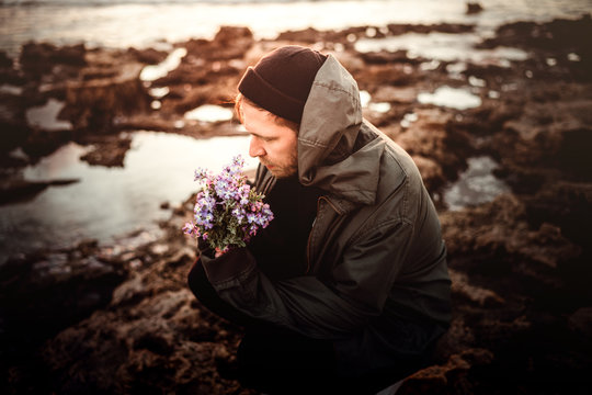 Junger Mann mit Jacke M&uuml;tze und Kapuze auf dem Kopf kniet an der Fels K&uuml;ste in Sizilien und hat Blumen in der Hand