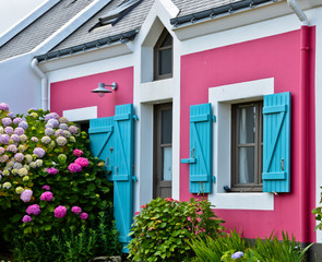 Fototapeta na wymiar Sonniger Blick auf eine Hausfront mit rosa Wand und leuchtend blauen Fensterläden. Davor blüht eine Hortensie.