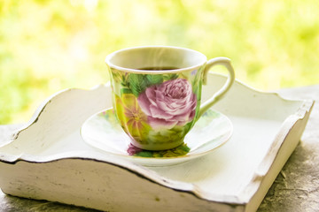 Hot morning coffee in small floral design cup on shabby chic vintage white tray on window sill in front of summer window
