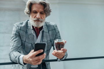 Serious bearded gentleman with drink using mobile phone