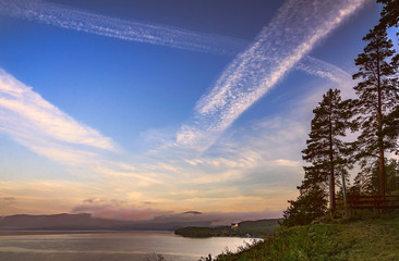 Picturesque landscape with a mountain lake, pine trees in the foreground, fog above the lake and among the mountains and a large sky with cirrus clouds