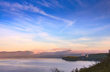 Bright colorful sunrise over a mountain lake. Blue sky with cirrus clouds, forested mountains in the distance and grass in the foreground