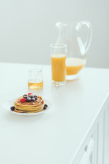 plate of pancakes with berries near glass and jug of orange juice and jar with honey on white table