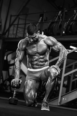 Fototapeta na wymiar Bodybuilder Posing in the Gym, Kneeling on the Floor With his hand on Dumbbell. Black and White Image