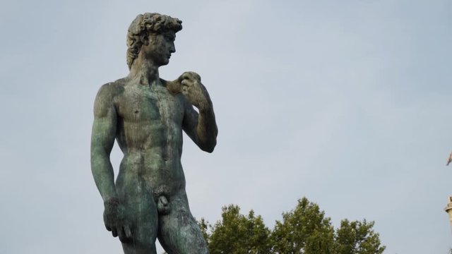Statue of David in park. Action. Beautiful manly statue of David stands in park on background of sky. Nude statue of David is made of dark stone and stands on pedestal in park