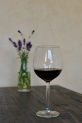 glass of red wine on a brown wooden table in front of light wall with green branch of  purple lavender in a vase