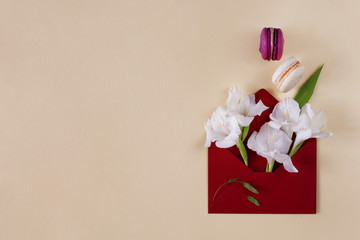 Creative minimalistic flower arrangement. White gladiolus and colorful macaroons in a dark red envelope on a beige pastel background. Minimalistic floral design, card, invitation. Flat lay, copy space