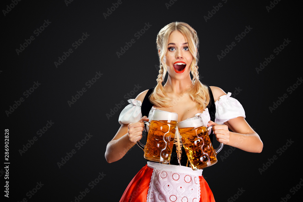 Wall mural Young sexy oktoberfest girl waitress, wearing a traditional Bavarian or german dirndl, serving two big beer mugs with drink isolated on black background.