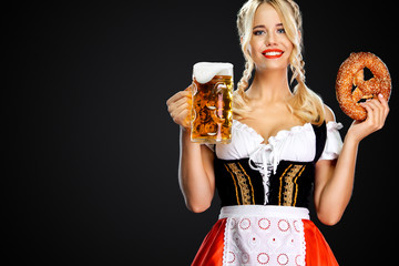 Smiling young sexy Oktoberfest girl waitress, wearing a traditional Bavarian or german dirndl, serving big beer mugs with drink and bretzel, isolated on black background.