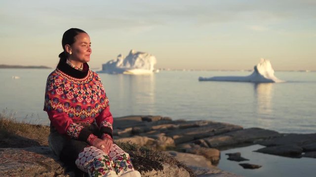 Woman in traditional dress looking at Disko Bay while sitting on rock against sky