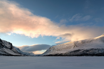 Tranquil winter scene of frozen lake Teusajaure in Lapland Sweden during calm morning with warm sunrise, lighted band of soft clouds, blue sky