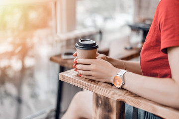 Paper coffee cup to go in woman's hands with red manicure while sitting in cafe.