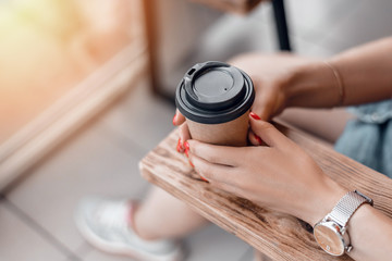 Paper coffee cup to go in woman's hands with red manicure while sitting in cafe.